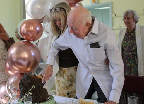 Old man in white shirt and blonde woman cutting cake together with pink balloons around them