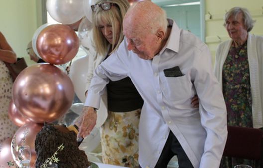Old man in white shirt and blonde woman cutting cake together with pink balloons around them