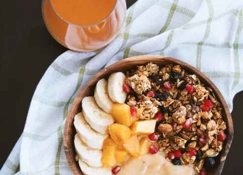 wooden bowl with banana and granola on top of green striped tea towel with glass of orange juice next to it