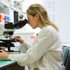 blonde woman in white lab coat looking through microscope