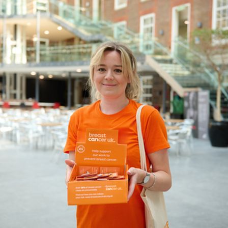 Blonde woman standing with orange charity collection box wearing orange breast cancer uk t-shirt