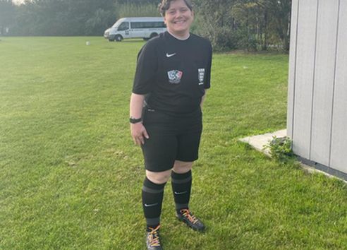 Person with brown hair in field in black sports kit and football boots with orange laces