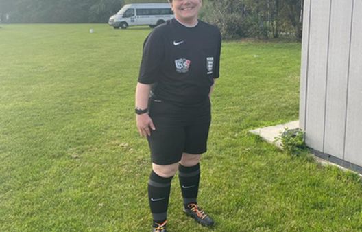 Person with brown hair in field in black sports kit and football boots with orange laces