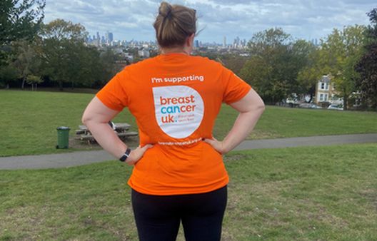 Woman with back to camera with orange Breast Cancer UK logo tshirt on in field