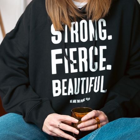 Woman with ginger hair wearing black jumper with white writing on holding a cup