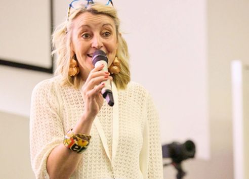 woman in cream jumper with blonde short hair and glasses on her head Talking into microphone in conference room