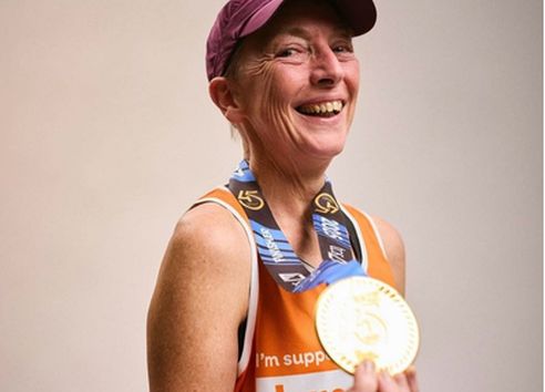Woman smiling headshot in orange running vest holding gold medal and wearing a purple cap