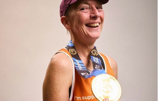 Woman smiling headshot in orange running vest holding gold medal and wearing a purple cap