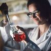Woman scentist in black framed glasses using pipette in glass container with red liquid