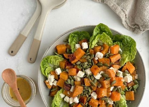 Sweet potato salad with feta and pumpkin seeds in a ceramic bowl next to ceramic serving spoons and a linen napkin
