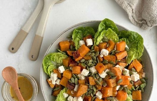 Sweet potato salad with feta and pumpkin seeds in a ceramic bowl next to ceramic serving spoons and a linen napkin