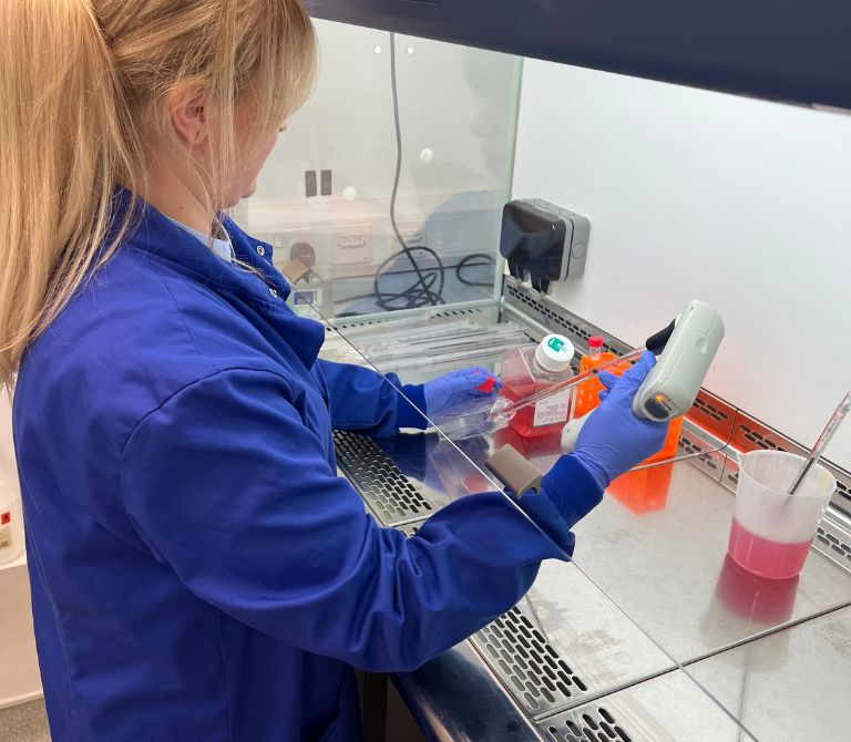 Woman wth blonde hair in blue lab coat putting something into petri dish