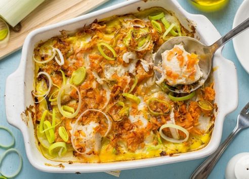 Baked leek, carrot, and hake gratin topped with golden cheddar cheese in a baking dish, being scooped with a stainless steel ladle.