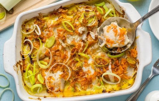 Baked leek, carrot, and hake gratin topped with golden cheddar cheese in a baking dish, being scooped with a stainless steel ladle.