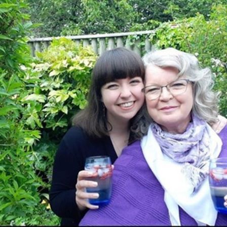 One young woman and one older woman hugging in front of trees wth cups of water in hands