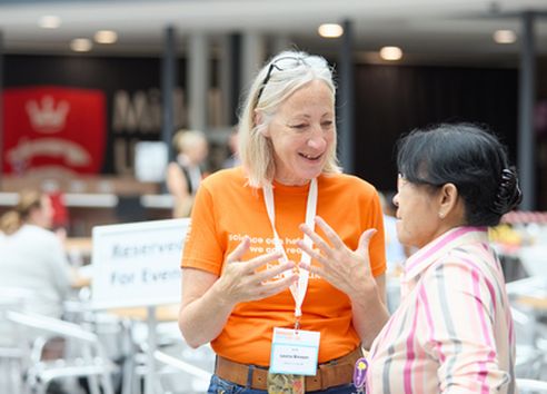 Woman with blonde hair in orange t-shirt speaking to shorter woman with black hair in pink striped shirt