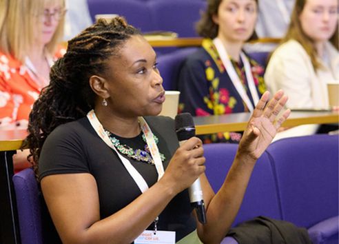 Woman in black t-shirt with white lanyard speaking in a lecture theatre sat down