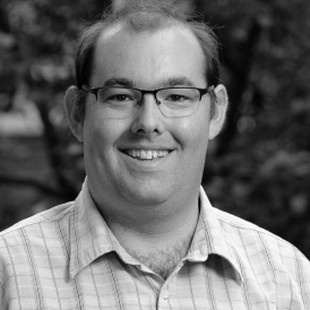 Man with thin brown hair in black and white head shot smiling at the camera in plaid shirt with glasses onDr Huw Jones