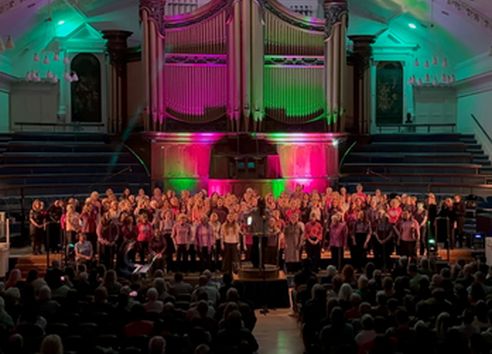 Group of people singing in choir in a church with pink and blue lights behind them