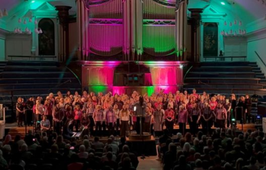 Group of people singing in choir in a church with pink and blue lights behind them