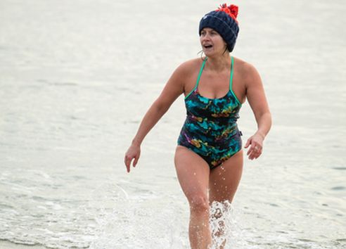 Woman in blue swimming costume and dark blue bobble hat walking out of the sea smiling
