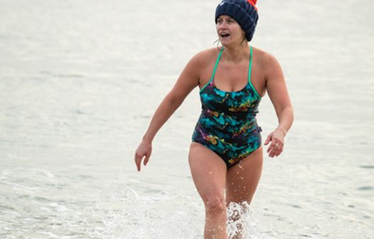 Woman in blue swimming costume and dark blue bobble hat walking out of the sea smiling