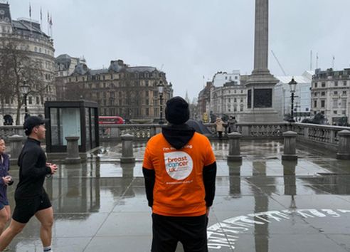 Man stood on pavement facing away from camera with black long sleeve t-shirt on with orange t-shirt on over the top with Breast Cancer Uk logo on