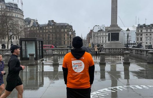 Man stood on pavement facing away from camera with black long sleeve t-shirt on with orange t-shirt on over the top with Breast Cancer Uk logo on