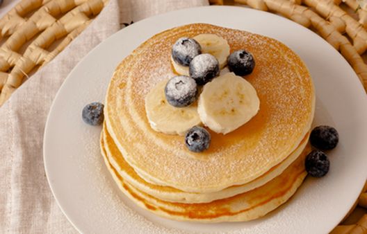 A stack of fluffy, healthy pancakes topped with fresh blueberries and banana, part of a Breast Cancer UK prevention-focused recipe guide.